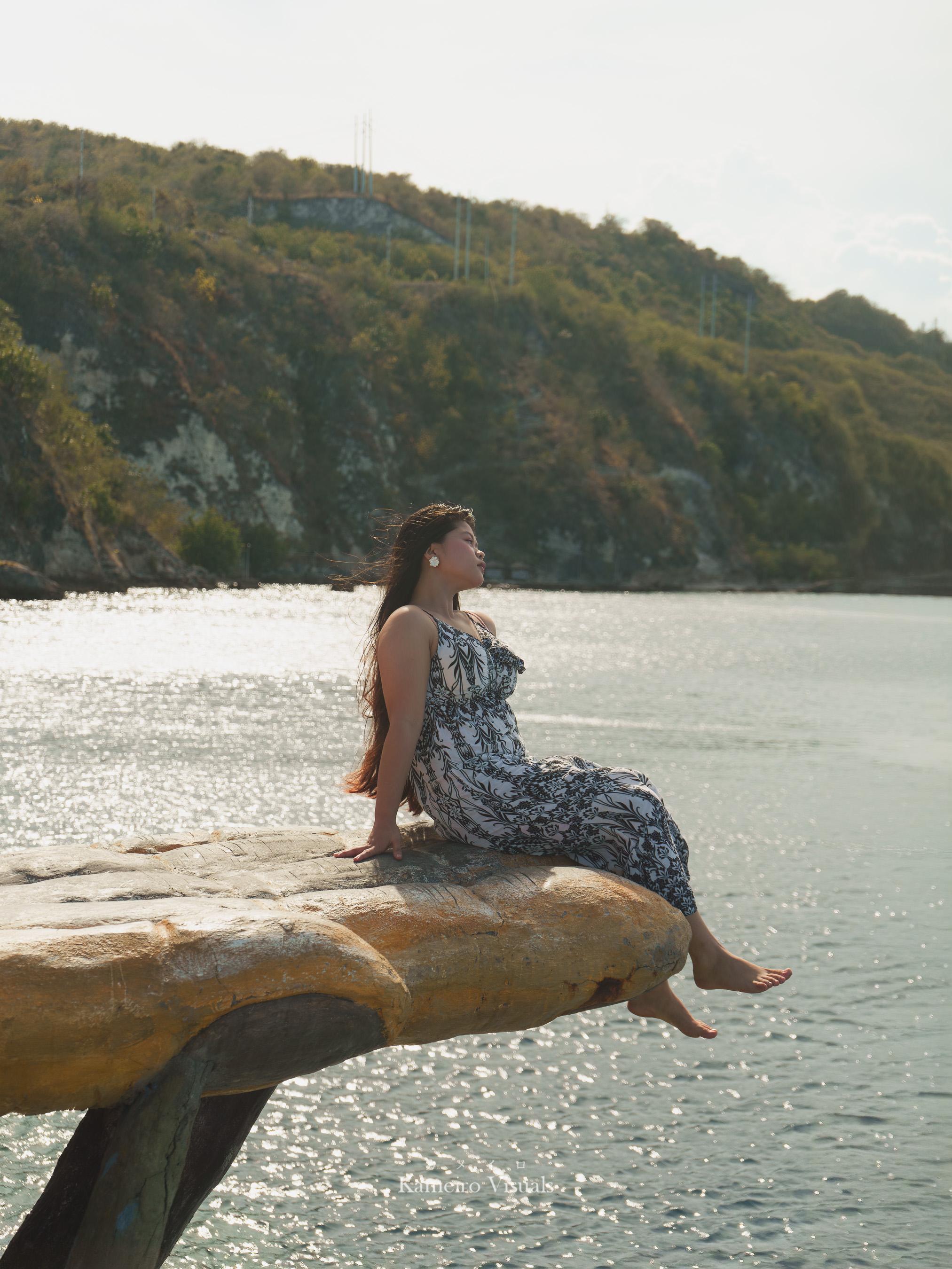 A woman seated on a rock beside sunlit water and a wooded hillside.