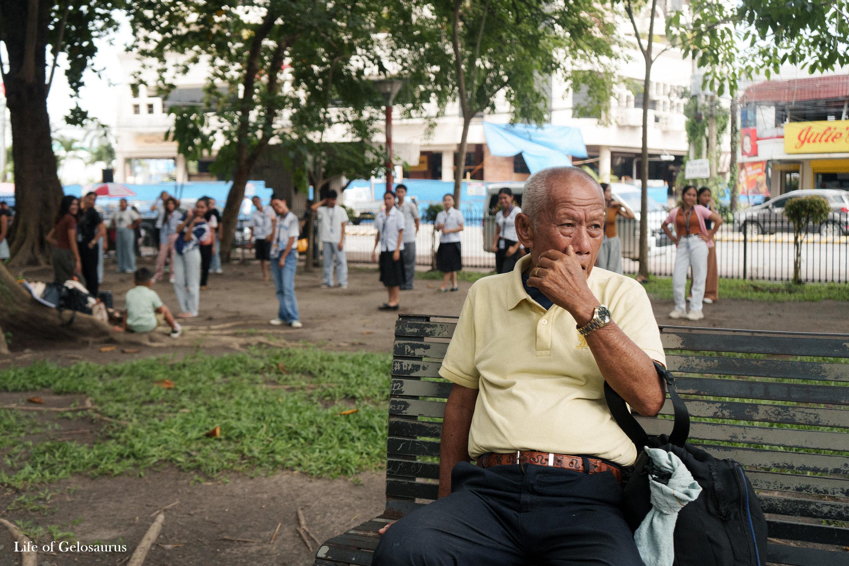 An older man seated on a bench in a park in General Santos City, with people and trees behind him.