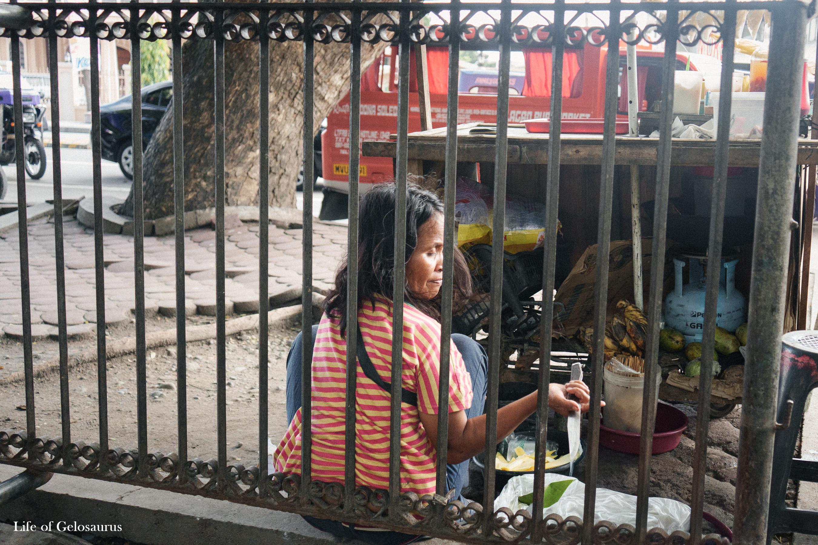 A woman seated behind a metal fence beside a roadside stall in General Santos City.