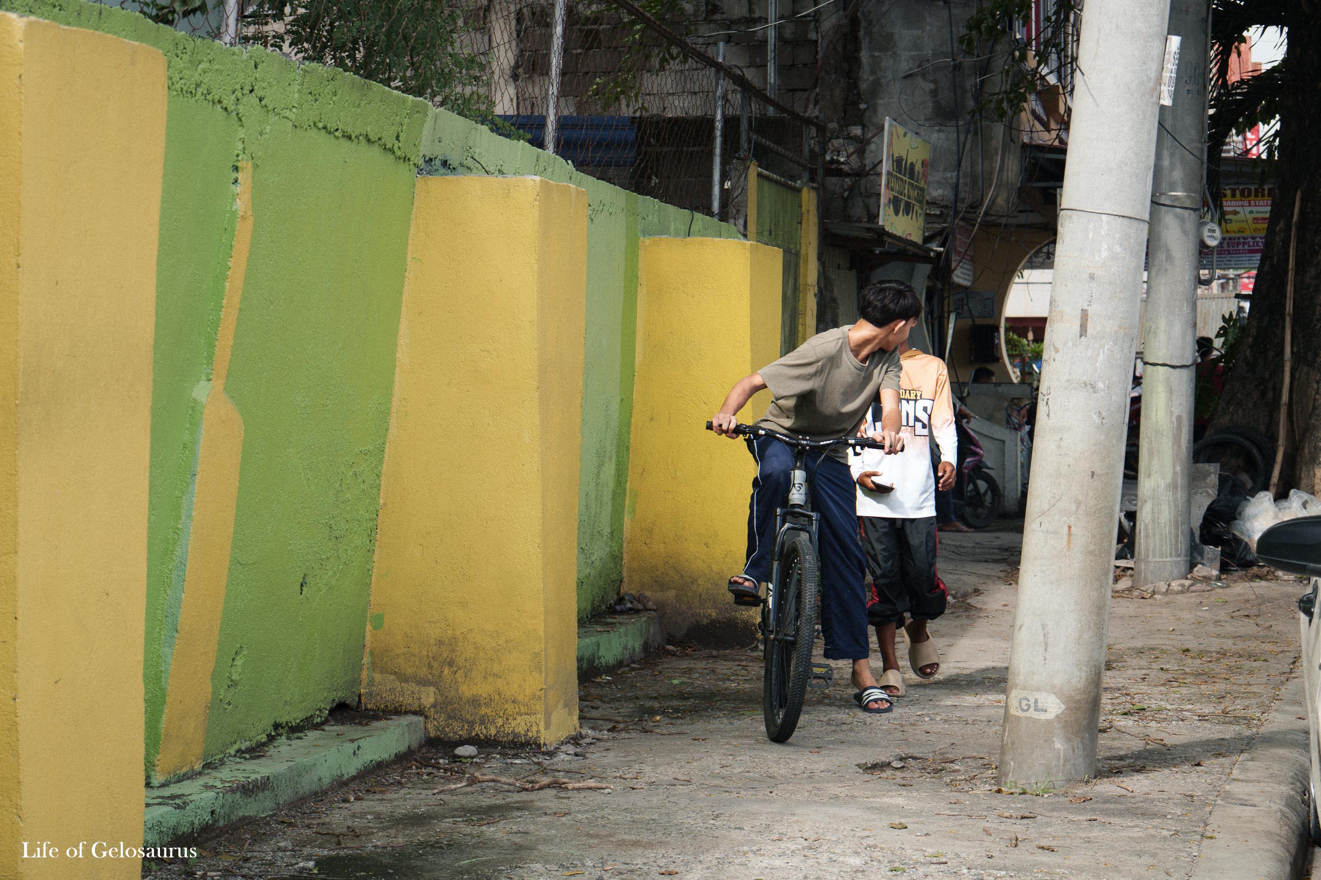Two boys riding a bicycle beside a green and yellow wall in General Santos City.